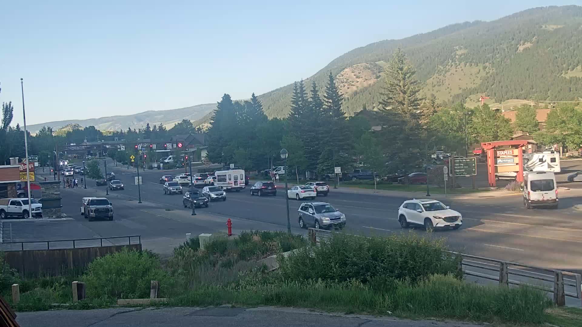 Moderate traffic flows along a multi-lane road in a town nestled in a valley beneath mountains on a clear, sunny day.