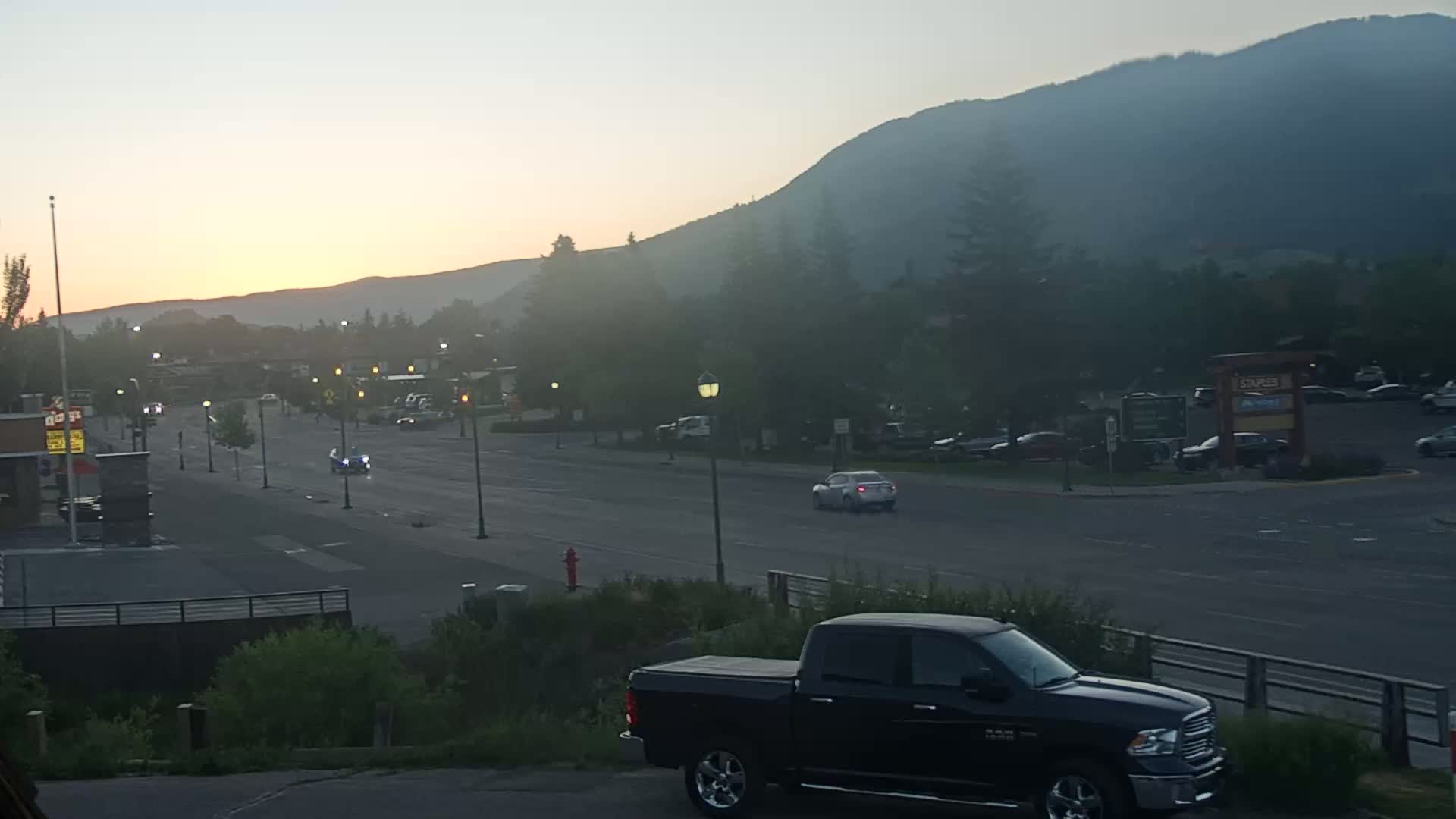 A hazy sunrise illuminates a street scene with several cars, a black pickup truck in the foreground, and mountains in the background.