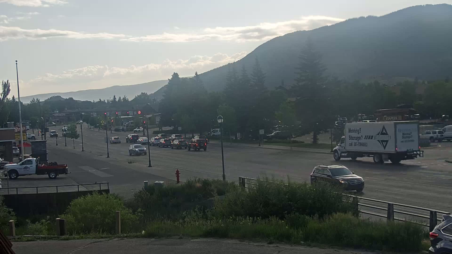 A hazy, partly cloudy day reveals a multi-lane road with moderate traffic in front of a mountain range, with some businesses and buildings visible along the roadside.