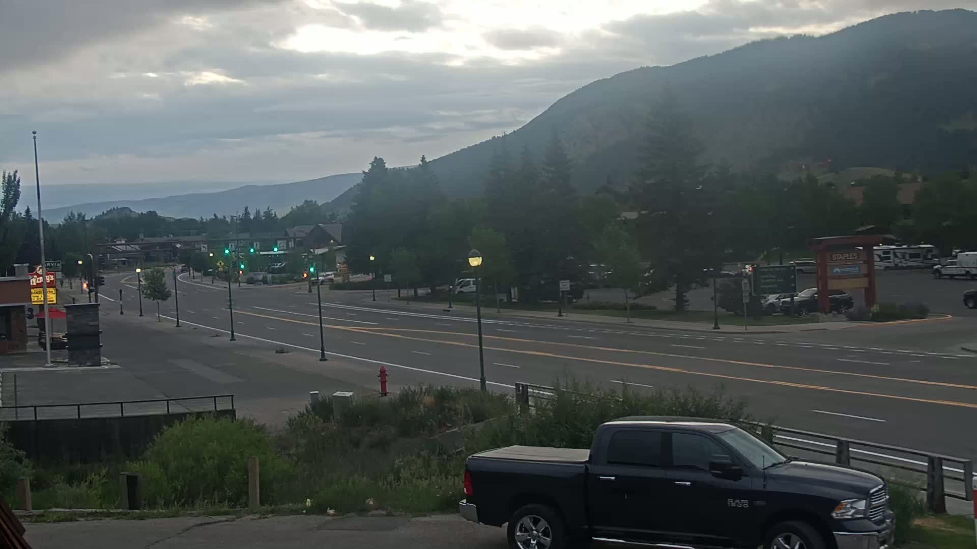 A mostly cloudy day reveals a long, straight road flanked by buildings and trees, leading toward a mountain range in the background.