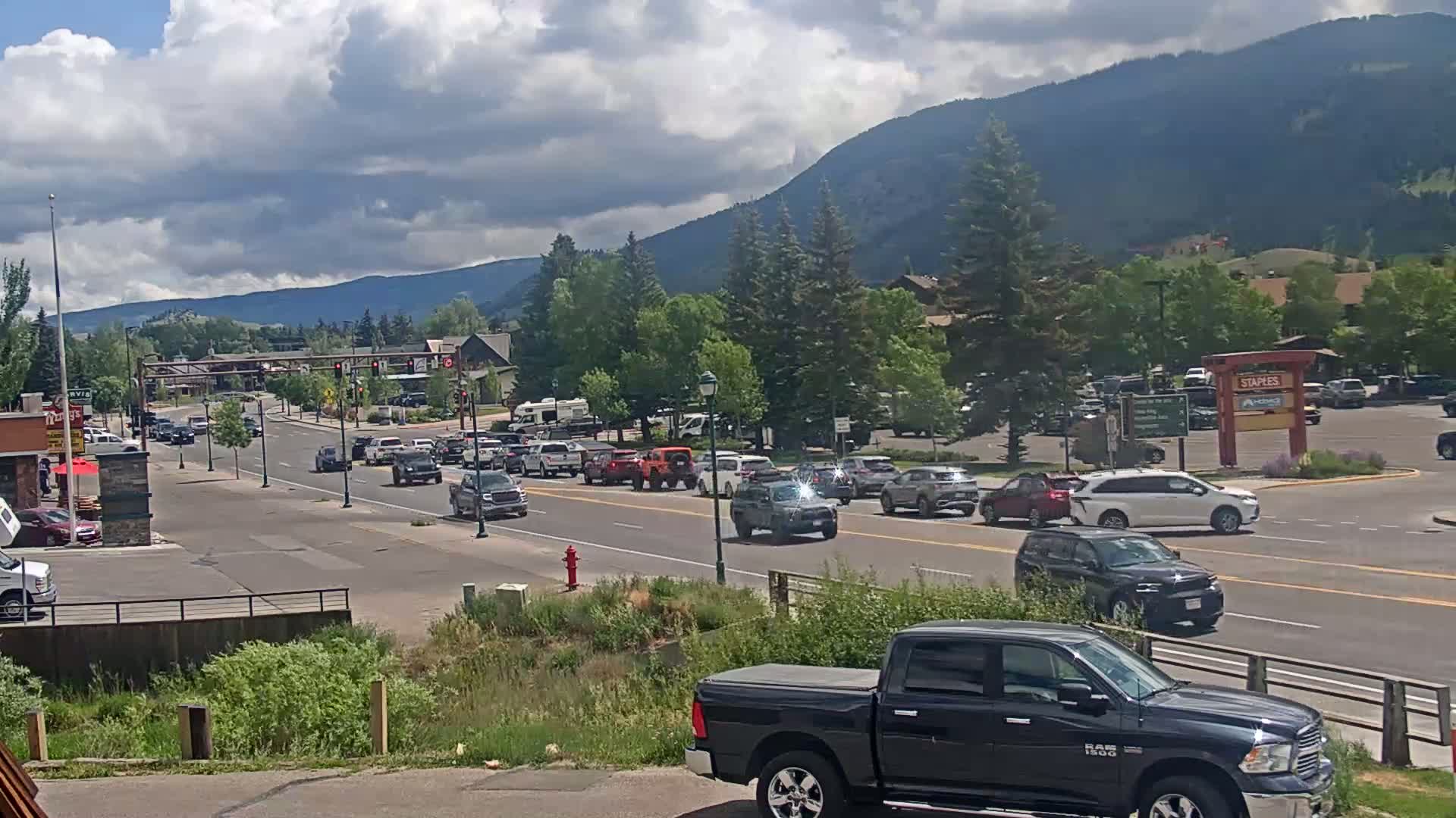 A partly cloudy day shows a busy street scene in a town nestled in a valley with mountains in the background.