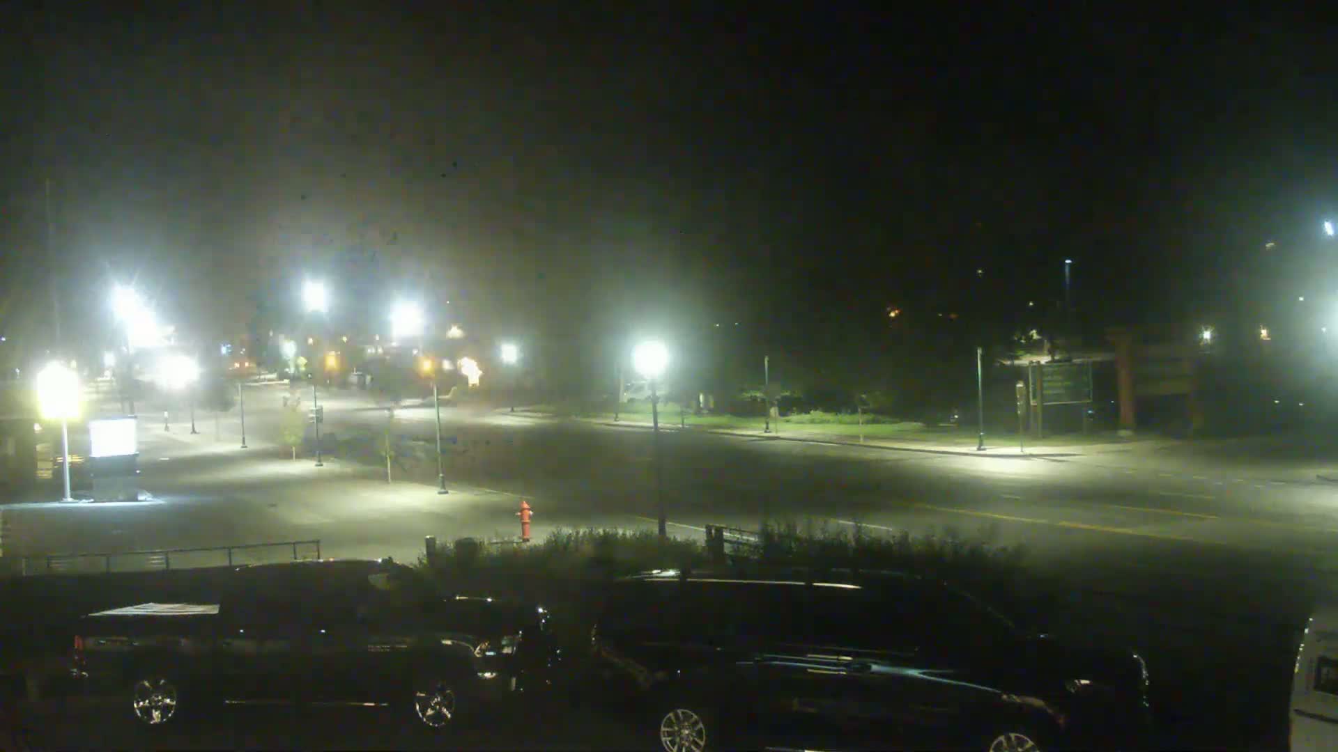 A nighttime scene shows a mostly empty street lit by lampposts, with several dark-colored vehicles parked in a lot, under hazy or foggy conditions.