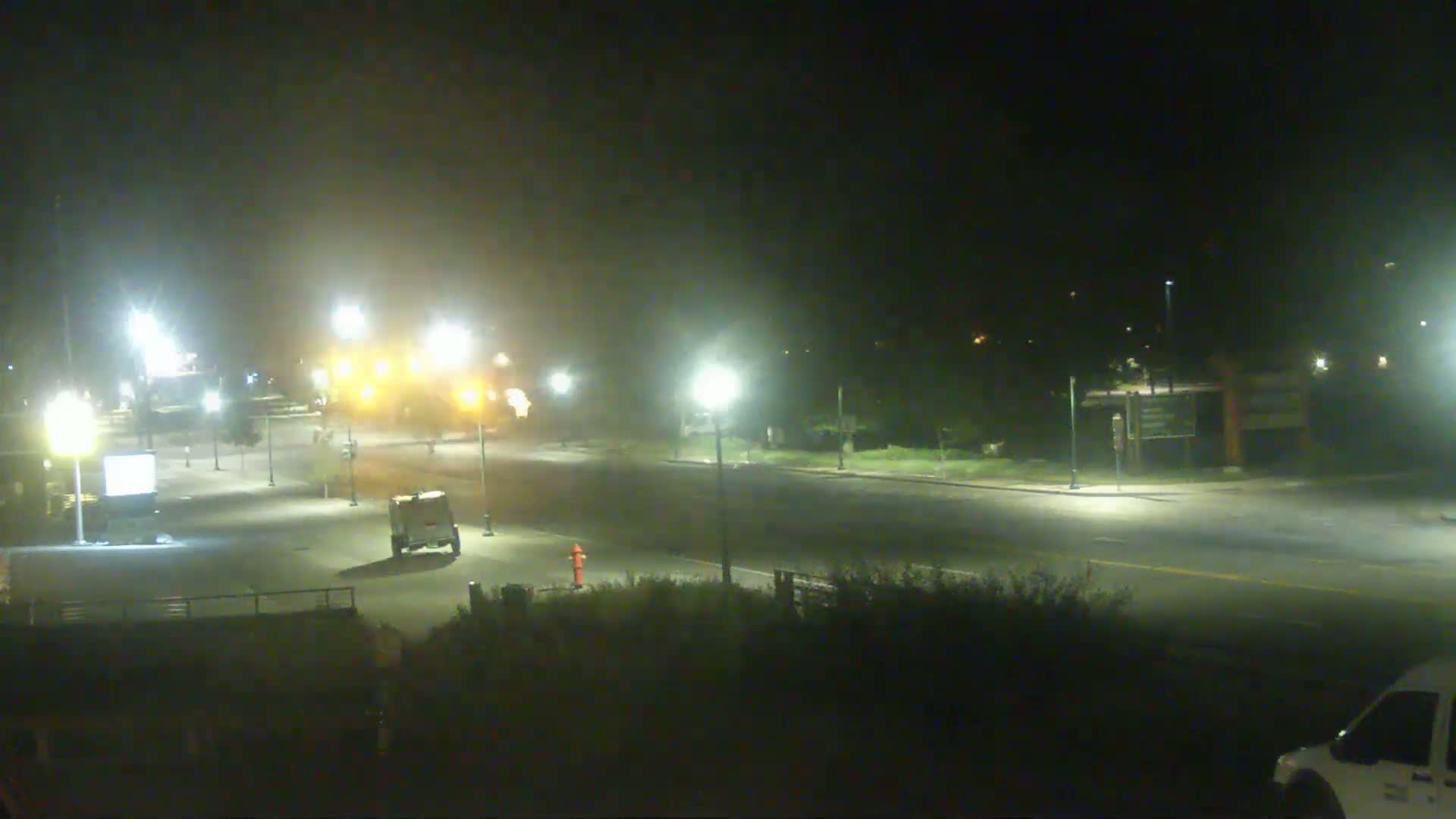 A hazy night scene shows a mostly empty road illuminated by streetlights, with a small vehicle parked near the intersection and a portion of a van visible in the bottom right corner.