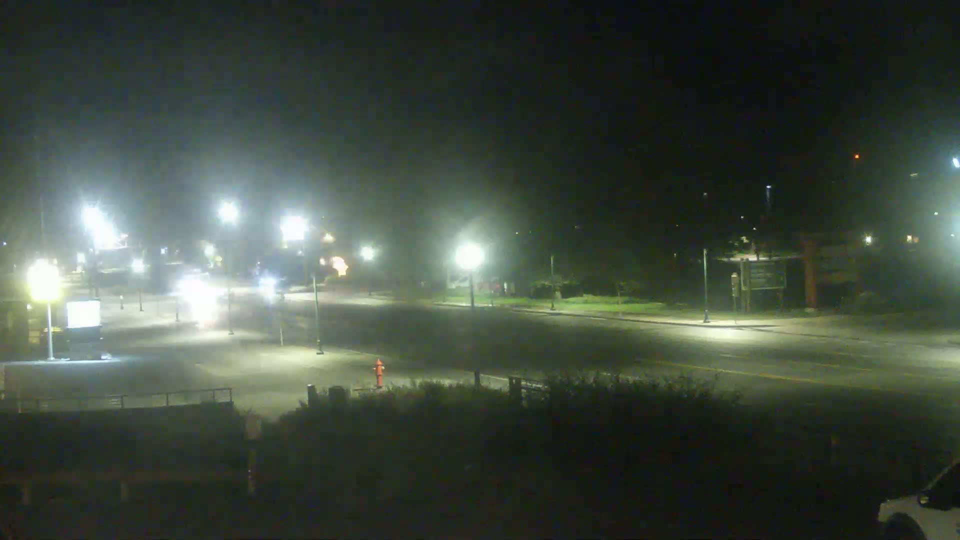 A nighttime street scene shows a mostly empty road with a few vehicles and streetlights under hazy conditions.