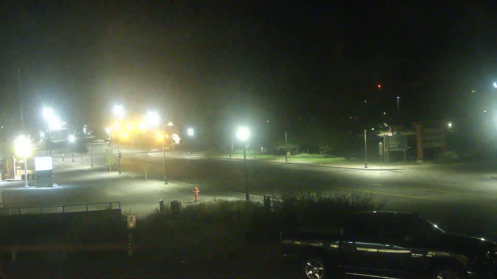 A hazy night scene shows a mostly empty road with streetlights illuminating a parking lot, some low vegetation, and a dark vehicle partially visible in the bottom right corner.