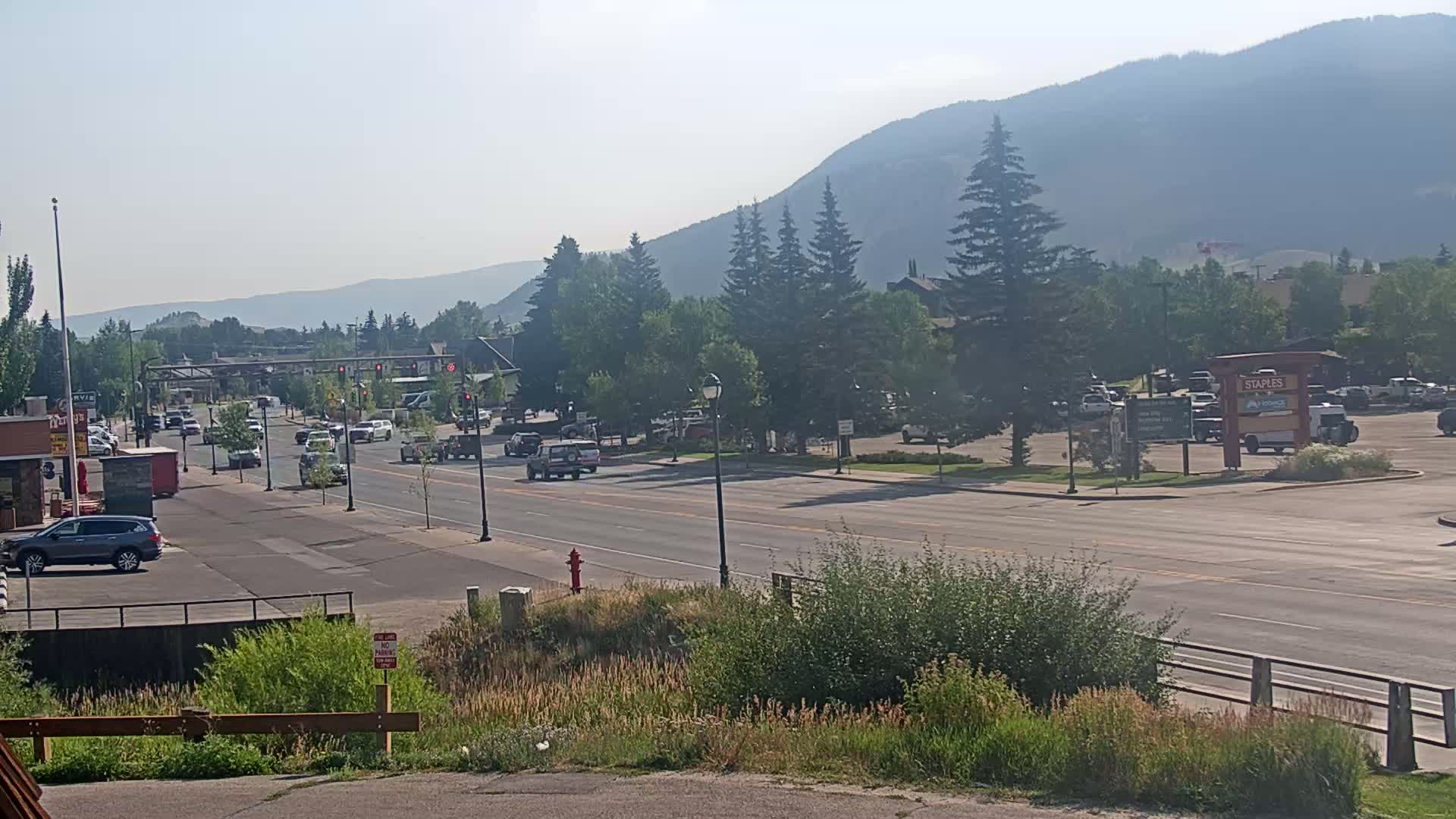 A hazy, sunny day reveals a multi-lane road with moderate traffic passing through a town nestled against a mountain range, lined with trees and businesses.