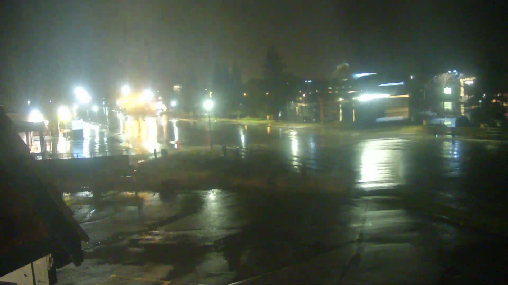 A hazy, rainy night view captures a wet outdoor urban area with numerous bright streetlights and building lights reflecting on the dark, water-slicked surfaces.