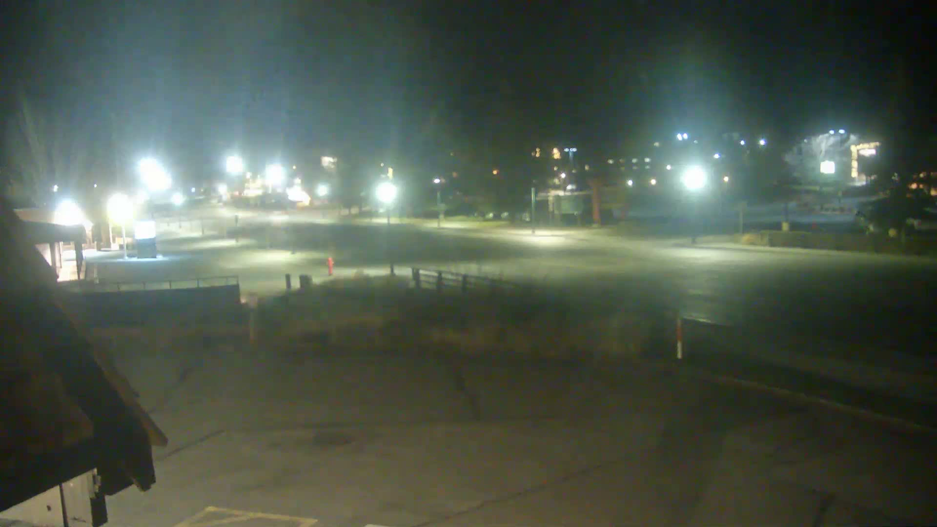A wide, outdoor nighttime scene reveals an empty paved area and grassy slopes illuminated by numerous streetlights and distant building lights under a clear sky, with a building roofline partially visible in the lower left.