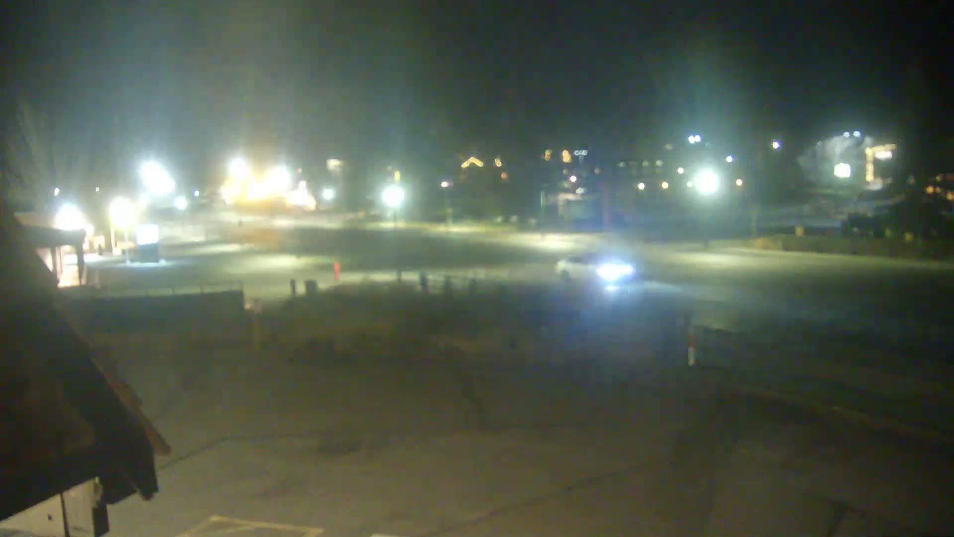 The image displays a clear night scene of an outdoor area, likely a parking lot or street, illuminated by numerous bright lights from buildings and streetlamps, with a car visible with its headlights on.