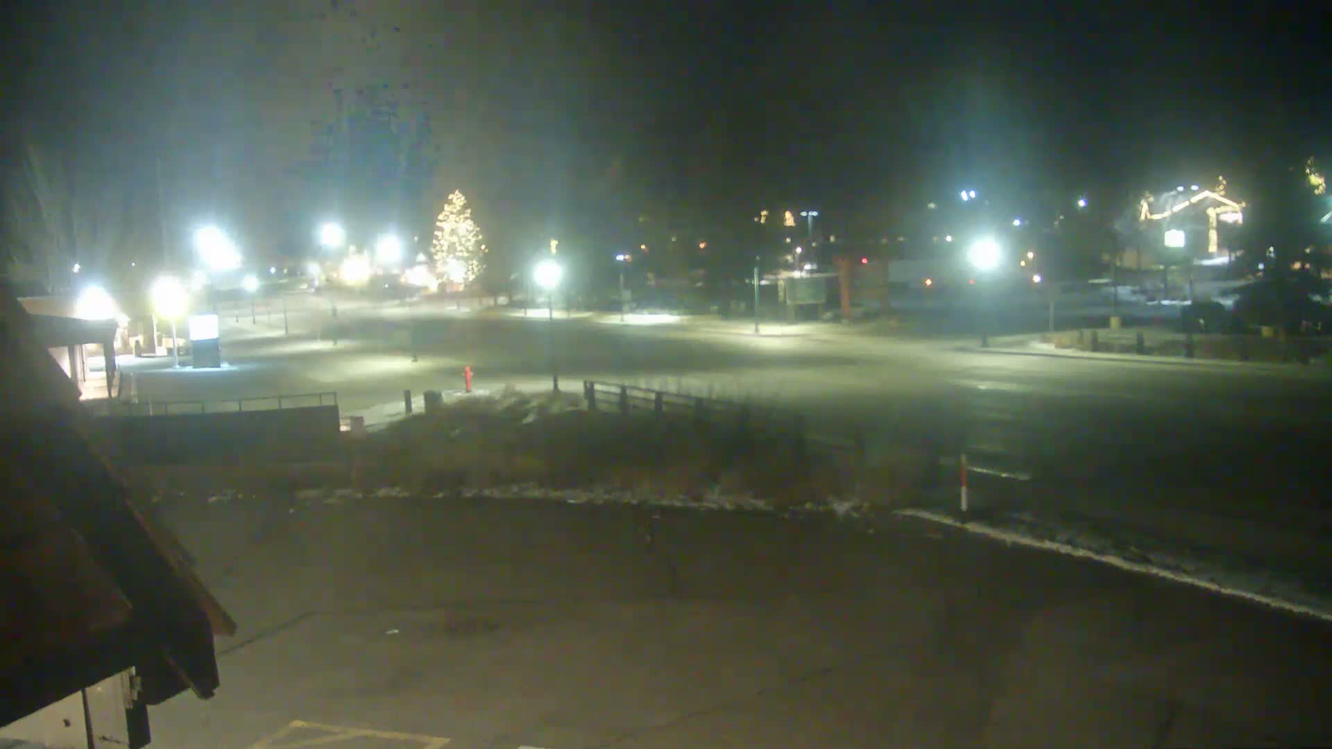 The image displays a quiet, hazy winter night scene in an outdoor area, featuring a brightly lit Christmas tree, numerous streetlights illuminating roads and patches of snow, and buildings adorned with holiday lights.