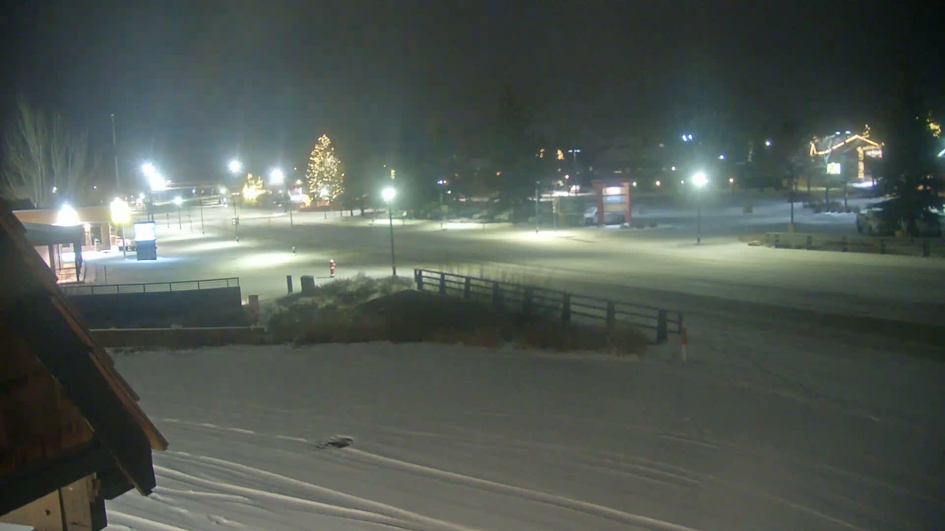 An empty, snow-covered outdoor area is brightly illuminated at night by numerous streetlights and festive holiday decorations on buildings and a large Christmas tree, under an overcast sky.
