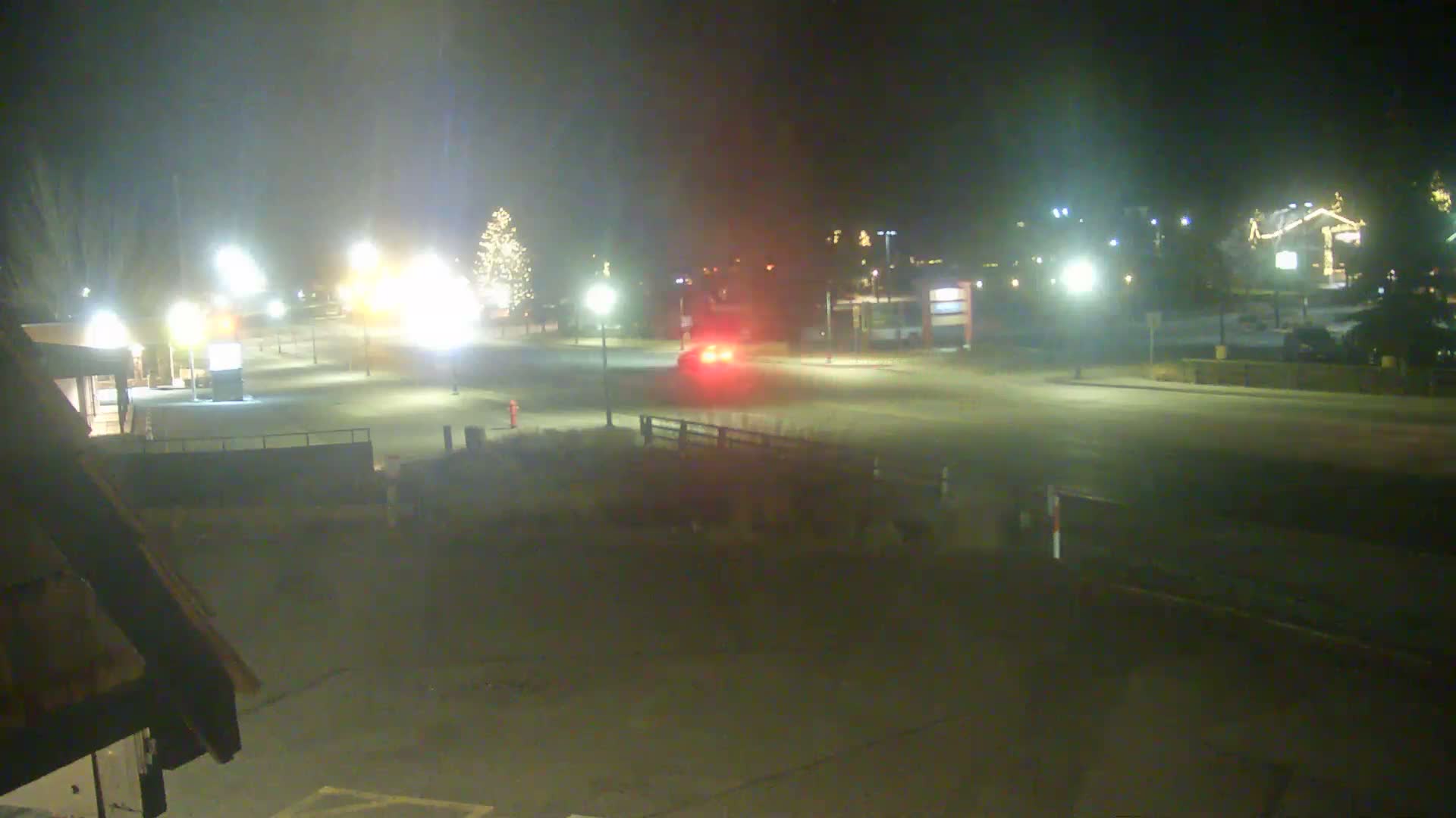 An empty, snow-covered outdoor area is brightly illuminated at night by numerous streetlights and festive holiday decorations on buildings and a large Christmas tree, under an overcast sky.