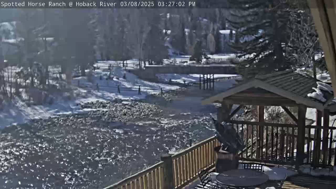 A partially snow-covered rocky river flows past a wooden deck with a gazebo, under a sunny sky.