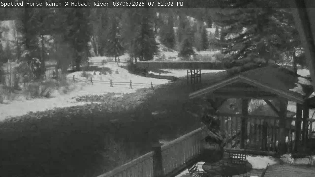 A snow-covered landscape is viewed from a wooden deck, overlooking a river that winds through a wooded area.