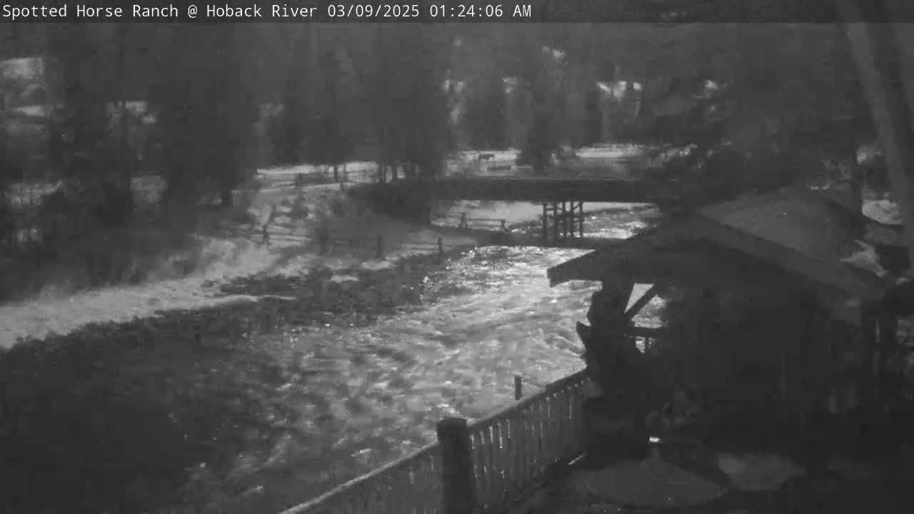 A nighttime, black and white view shows a swiftly flowing river under a bridge, next to a wooden structure and fence.