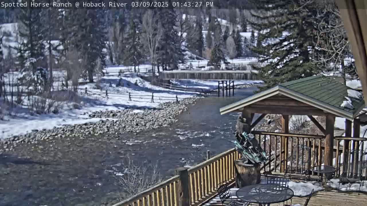 A partially snow-covered river flows past a wooden deck with a gazebo, under a sunny sky.