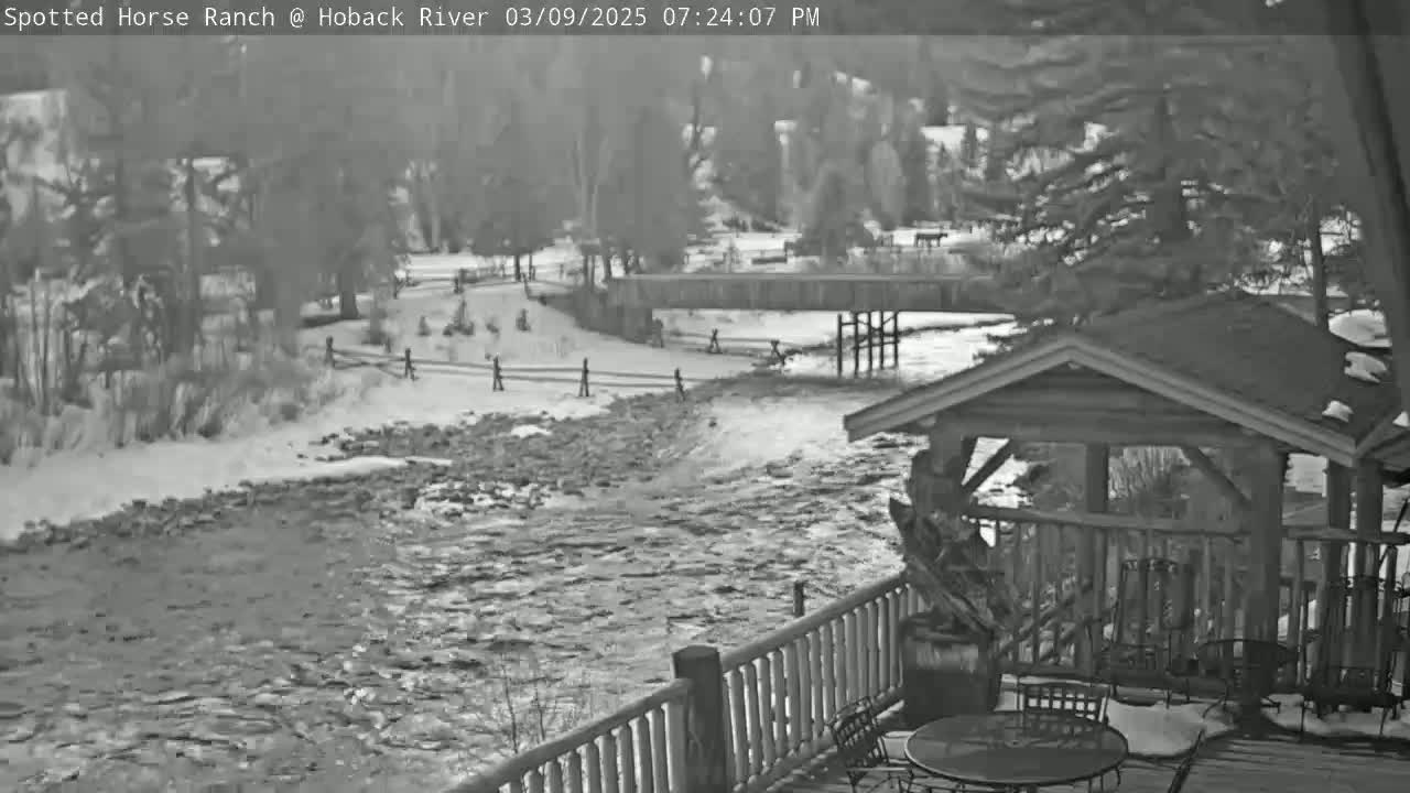 A snow-covered river flows past a wooden bridge and a rustic outdoor seating area with a gazebo.