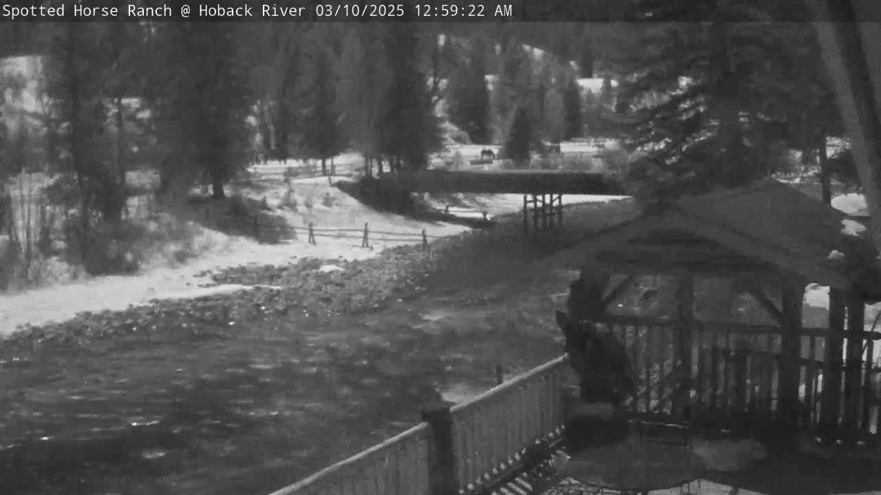 A grayscale image shows a flowing river next to a snow-covered bank, with a wooden gazebo and railing partially visible in the foreground under overcast skies.