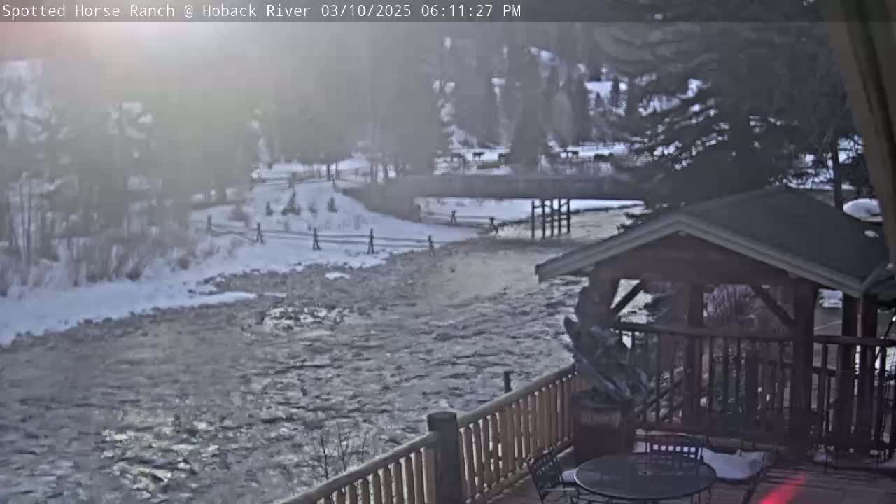 A view from a deck overlooking a swiftly flowing river partially covered in ice, with snow on the banks and trees in the background on an overcast day.