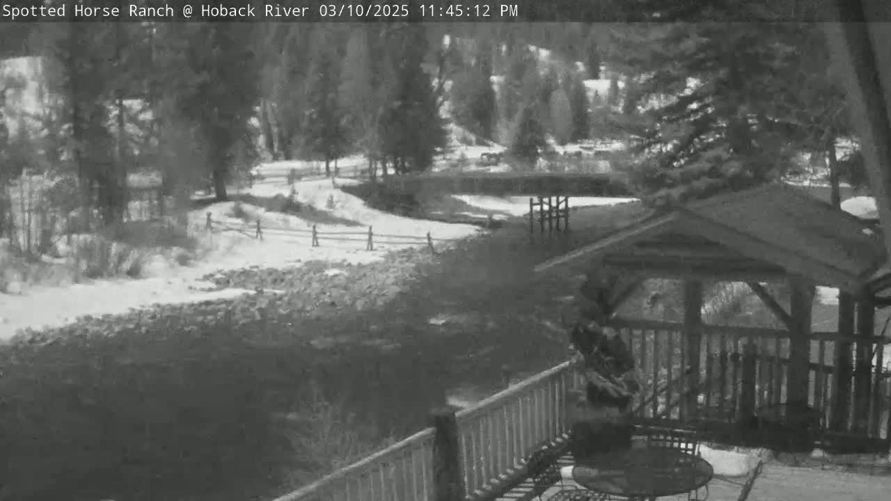 A snow-covered landscape with a river, wooden fences, trees, and a gazebo viewed from a deck.