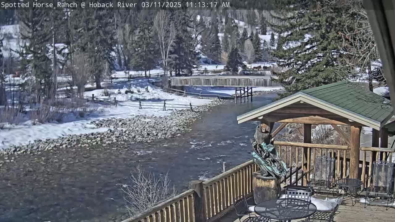 A sunny, snow-dusted riverbank scene features a flowing river, a wooden footbridge, and a partially visible outdoor patio with a gazebo under a clear sky.
