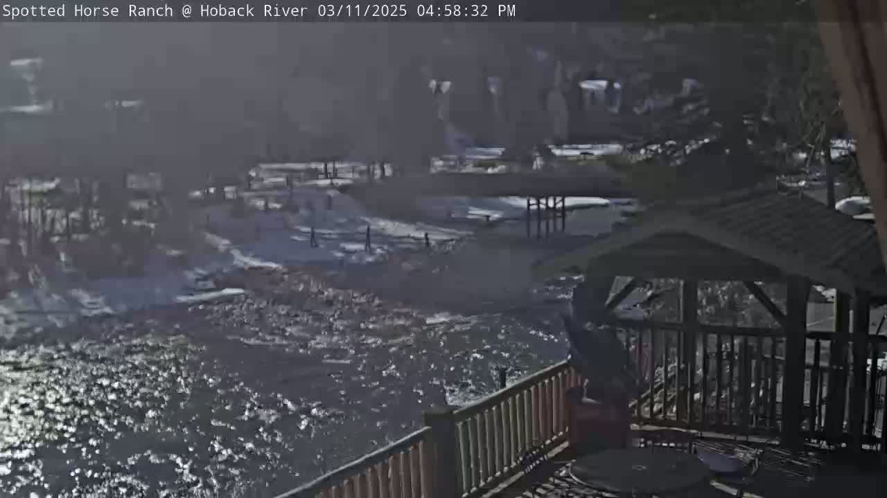 A partially sunny view overlooking a swiftly flowing river, with a wooden gazebo and deck in the foreground and a snow-dusted landscape in the background.