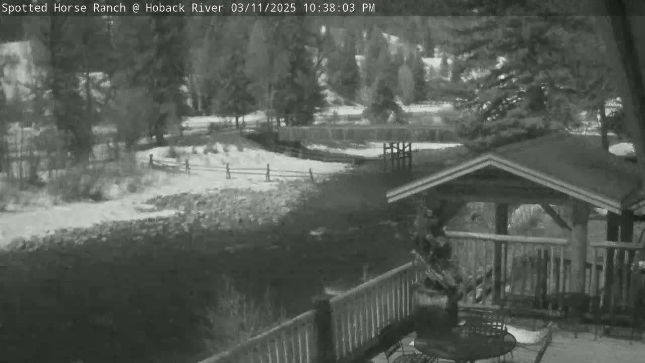 A snow-covered riverbank with trees and a wooden structure with a deck overlooking a dark river under an overcast sky.