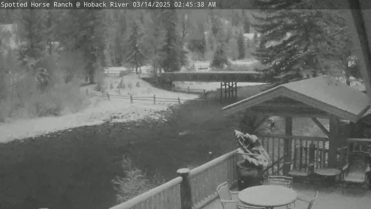 A snow-covered outdoor scene shows a wooden deck with patio furniture overlooking a snow-dusted riverbank and wooded area.