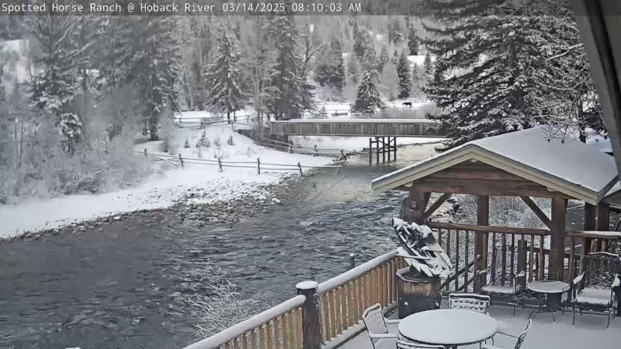 A snow-covered river flows beneath a wooden bridge, with a snow-dusted patio and wooden gazebo in the foreground, all surrounded by snow-laden trees.