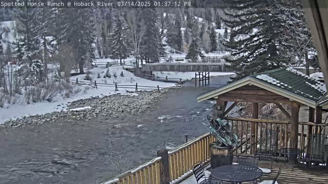 A snow-covered river flows beneath a wooden bridge, viewed from a deck with a wooden gazebo and patio furniture.