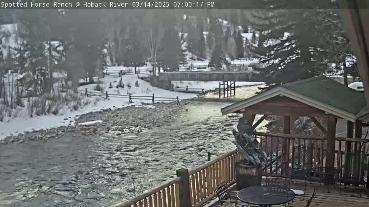 A partially snowy landscape shows a rushing river flowing under a bridge, viewed from a wooden deck with a gazebo and a sculpture.