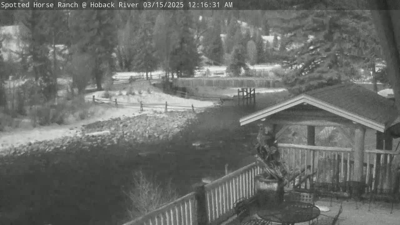 A snow-dusted riverbank with a wooden deck and cabin in the foreground, overlooking a calm river under a cloudy sky.