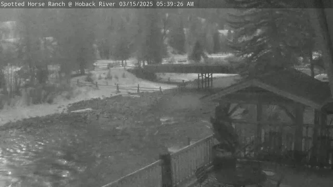 A snow-covered area with a swollen river flows past a wooden structure and a fence, under an overcast sky.
