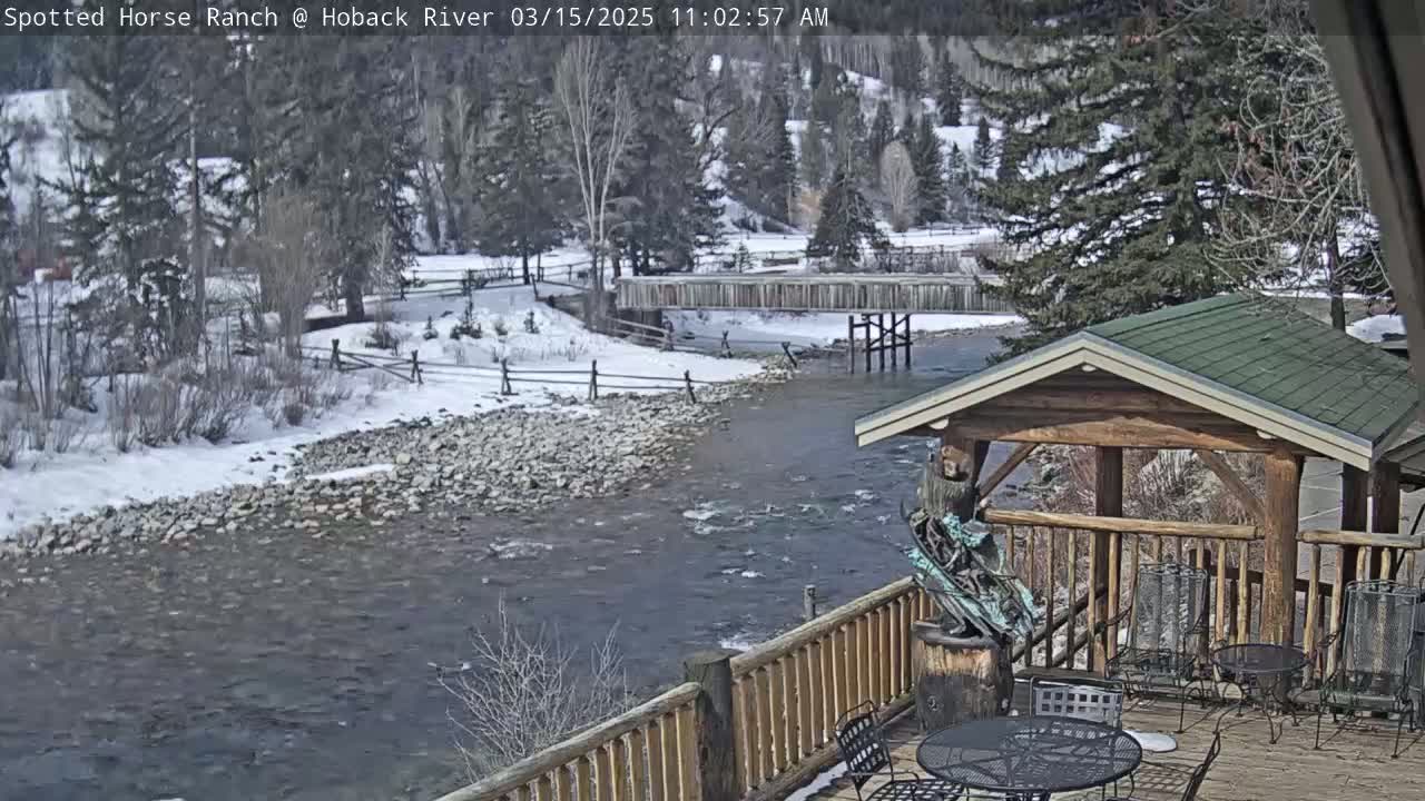A snow-covered riverbank with a wooden bridge and a wooden deck with outdoor furniture is seen from a deck overlooking a flowing river.