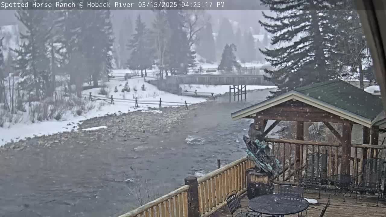 A snow-covered, rocky river flows past a wooden deck with a gazebo, under a lightly snowing sky.