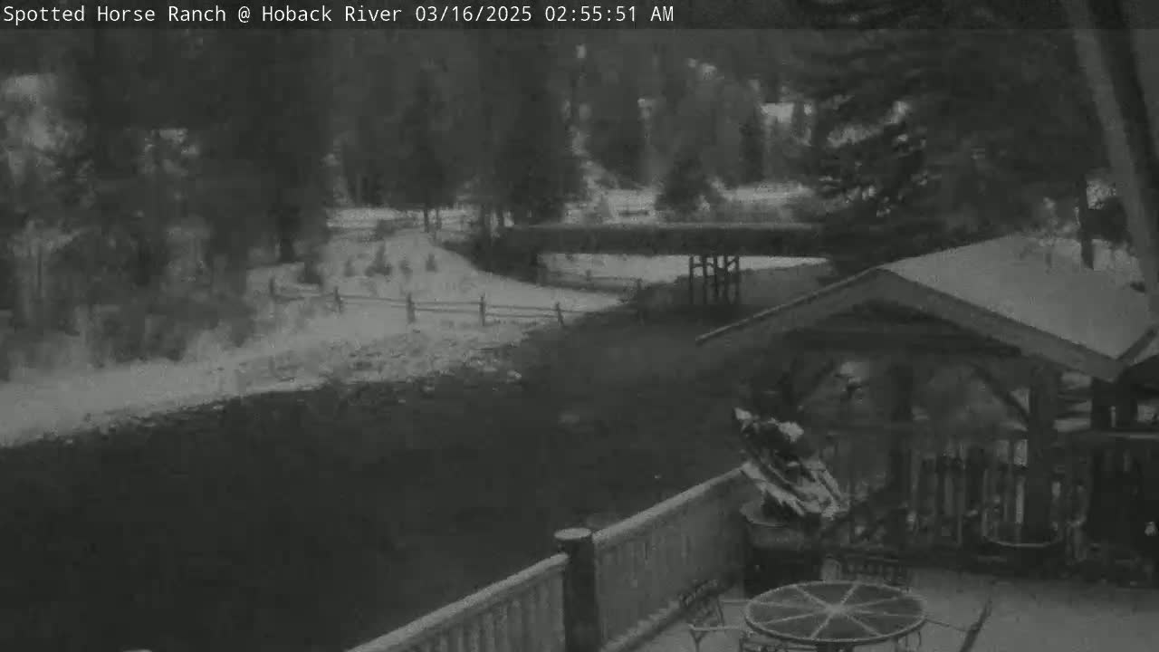 A nighttime, snowy scene shows a wooden deck with a table and chairs overlooking a snow-covered path, fence, and bridge, beyond which are dark trees.