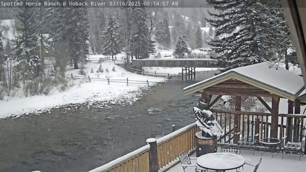 A snow-covered wooden deck overlooks a river flowing under a bridge, with snow-covered trees and a snow-covered landscape in the background during a snowfall.