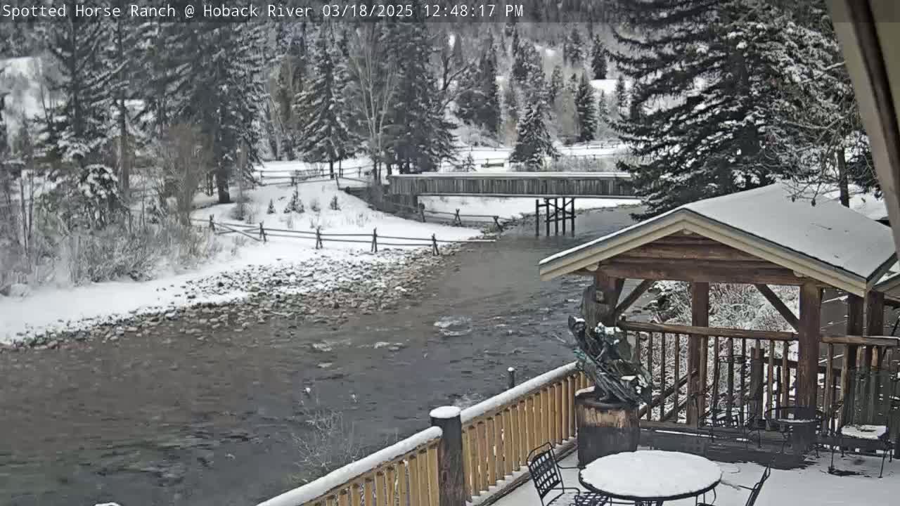 A snow-covered river flows under a wooden bridge, with a snow-dusted wooden gazebo and deck in the foreground, surrounded by snow-covered trees.