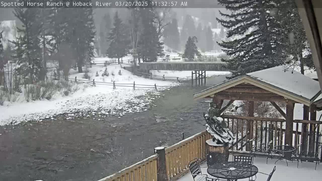 A snow-covered wooden deck overlooks a river flowing under a bridge, with snow falling and trees lining the riverbanks.