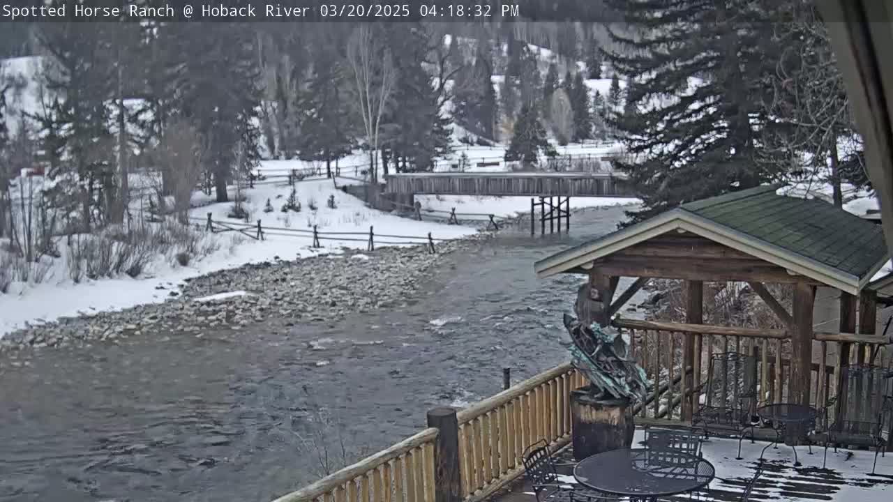 A snow-covered river flows past a wooden deck with a gazebo and metal chairs, under a partly cloudy sky.
