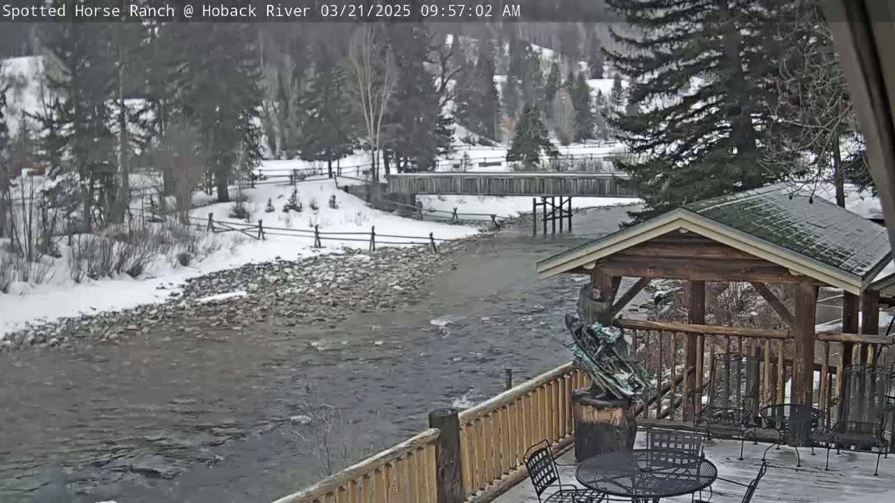 A snow-covered river flows beneath a wooden bridge, past a rocky bank, toward a partially visible wooden deck with outdoor furniture and a sculpture.