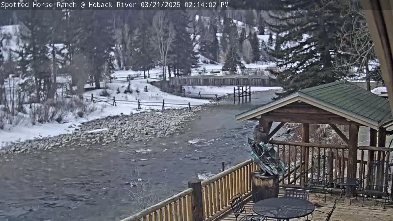 A snow-covered river flows past a wooden deck with a gazebo and outdoor seating, under a partly cloudy sky.