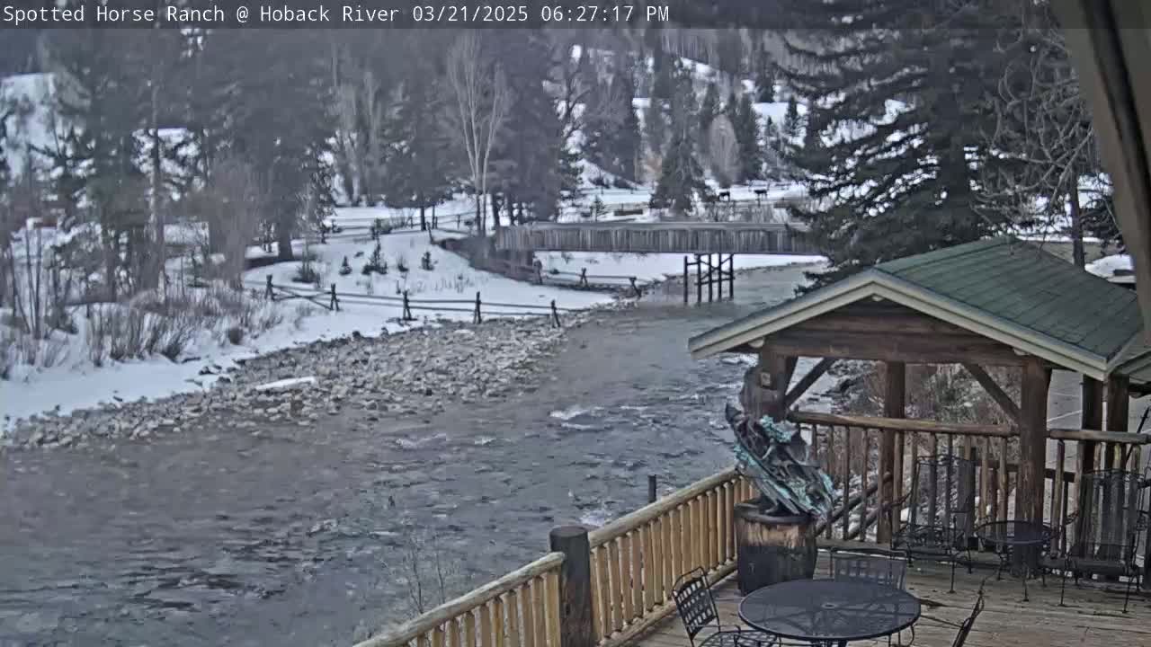 A partially frozen river flows past a wooden deck with a gazebo, under a partly cloudy sky.