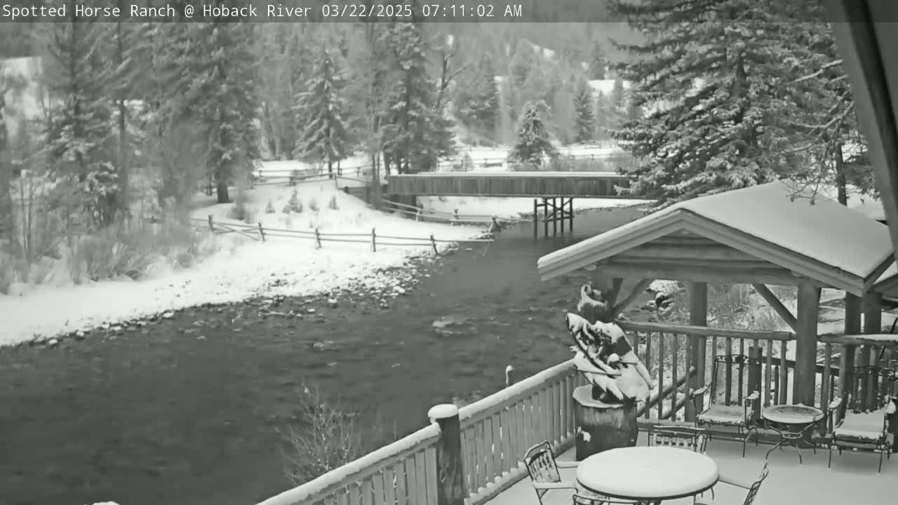 A snow-covered deck with outdoor furniture overlooks a river, a wooden bridge, and snow-covered trees.