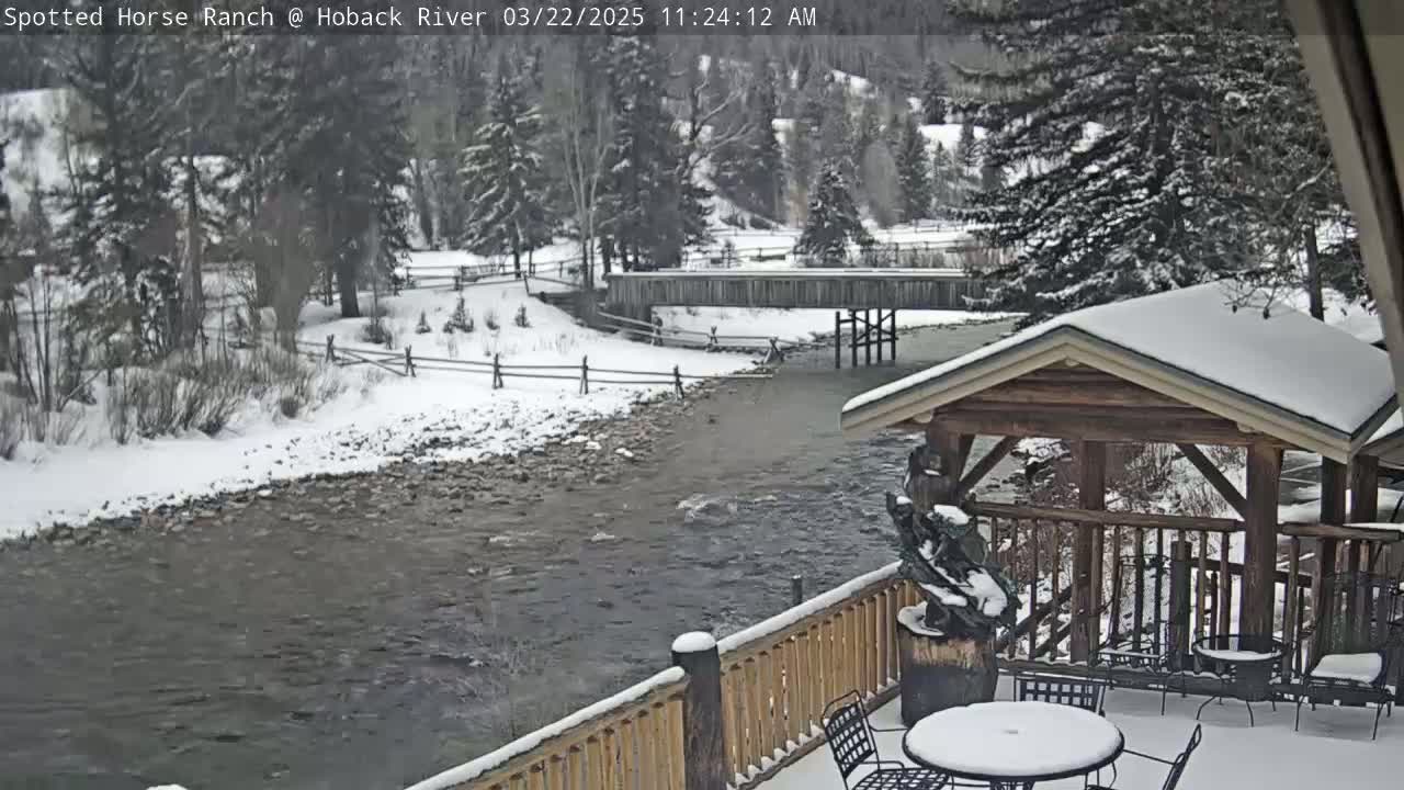 A snow-covered river flows beneath a wooden bridge and beside a snow-covered deck with a gazebo, tables, and chairs.