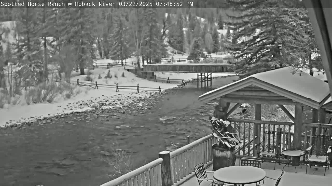 A snow-covered deck with outdoor furniture overlooks a river flowing under a wooden bridge, with snow-covered trees and a wooden fence in the background.