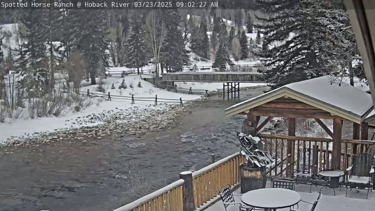 A snow-covered river flows beneath a wooden bridge and alongside a deck with a gazebo, all surrounded by snow-covered trees and a snowy landscape.