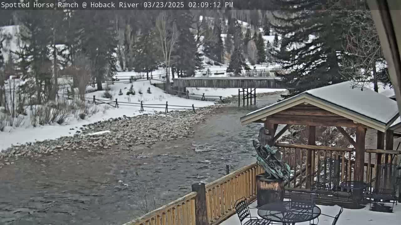 A snow-covered river flows under a wooden bridge, viewed from a wooden deck with a gazebo and outdoor furniture in the snowy landscape.