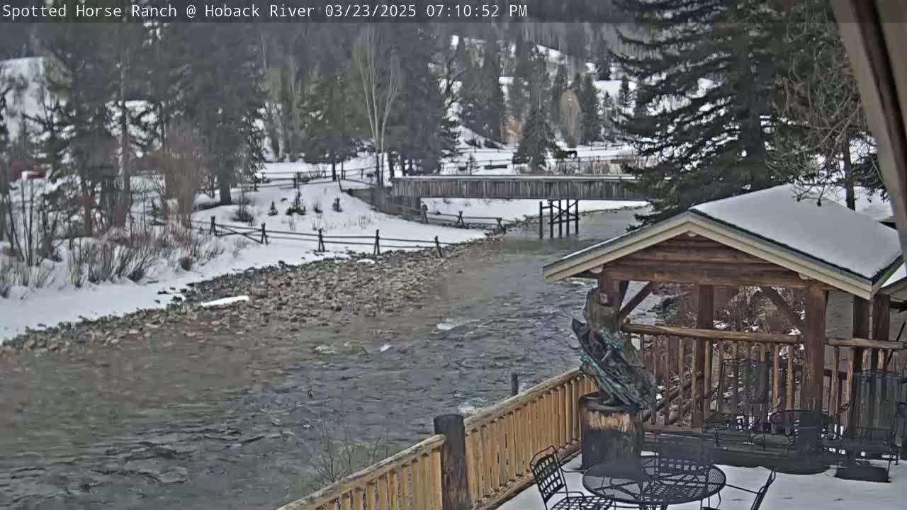 A snow-covered river flows under a wooden bridge, past a snow-dusted wooden gazebo with a metal sculpture, and alongside a snow-covered riverbank with trees in the background.
