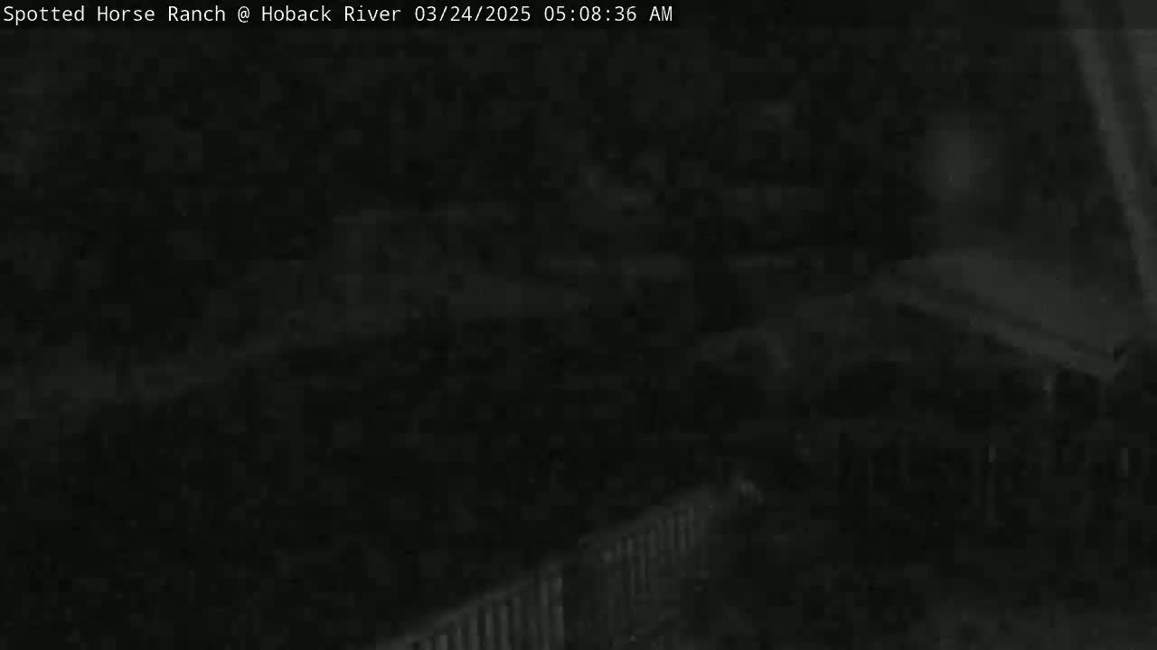 A dark, nighttime image shows an indistinct outdoor scene with a fence and what appears to be a building or structures in the background.